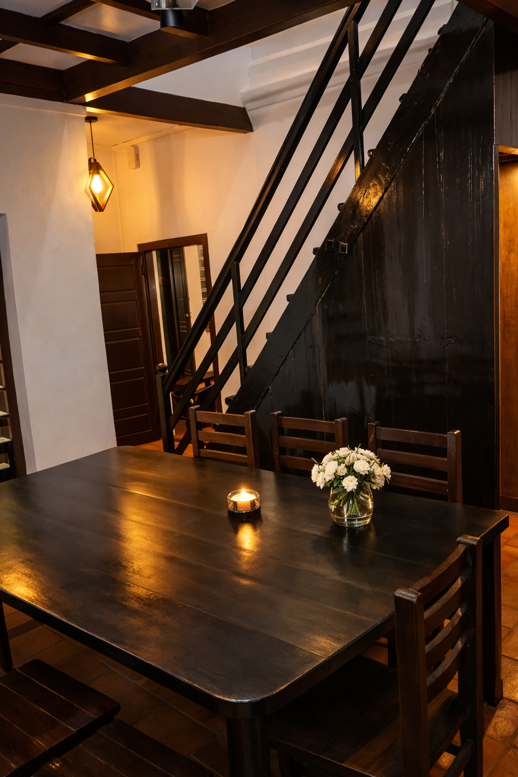 Rustic dining room with dark wood table, black staircase, candle, and white flowers in a vase.