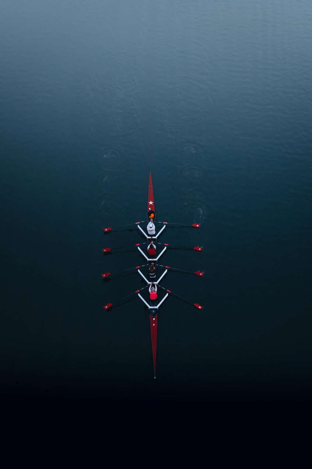 Aerial view of a four-person rowing crew in a red scull boat gliding on calm dark blue water.