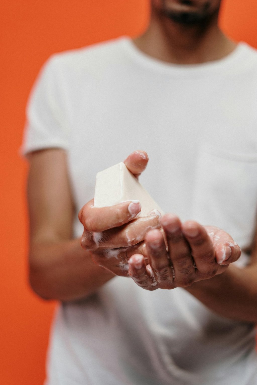 A man washing his hands with a bar of soap and white foam against an orange background.