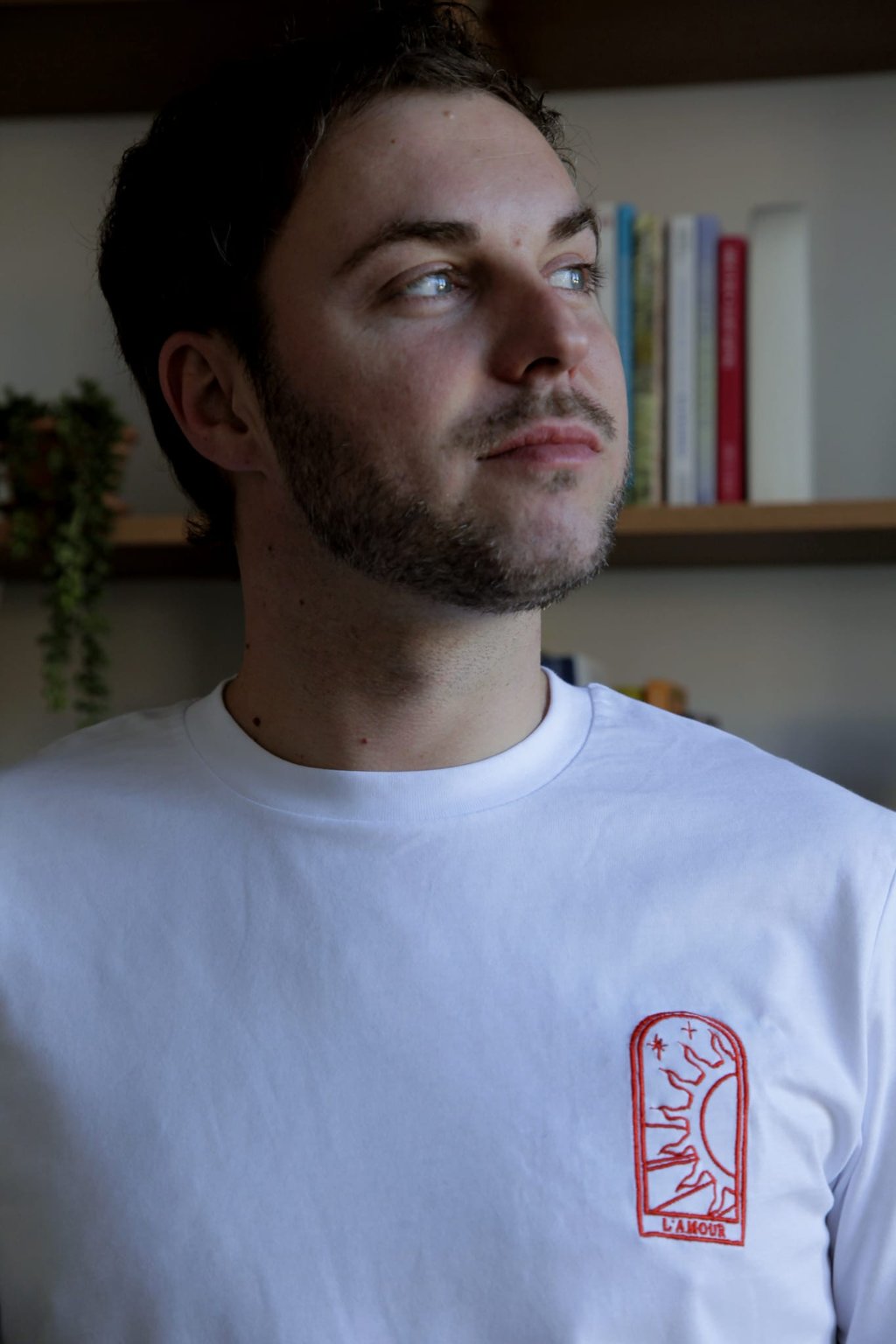 Young man with a beard wearing a white t-shirt featuring a red embroidered L'Amour sun graphic.