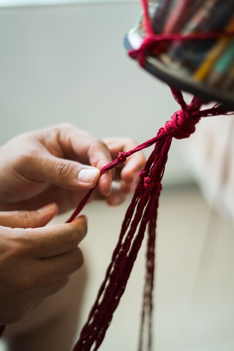 Manos tejiendo maceta de macrame color rojo con madera natural
