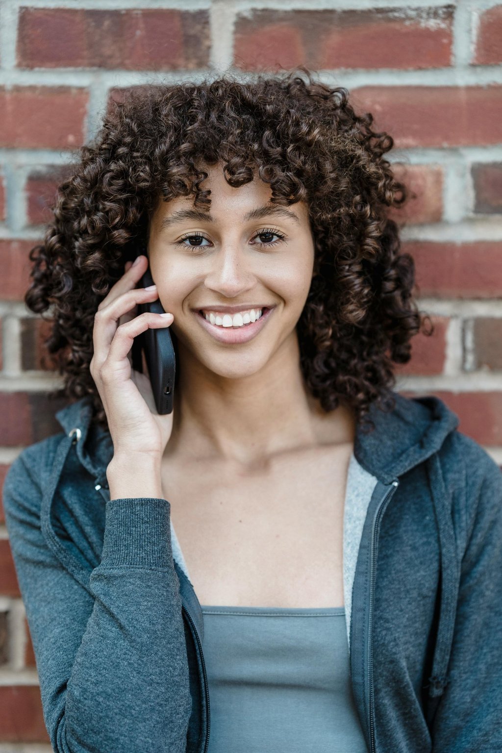 a woman with curly hair and a gray jacket. She's smiling and talking on the phone.