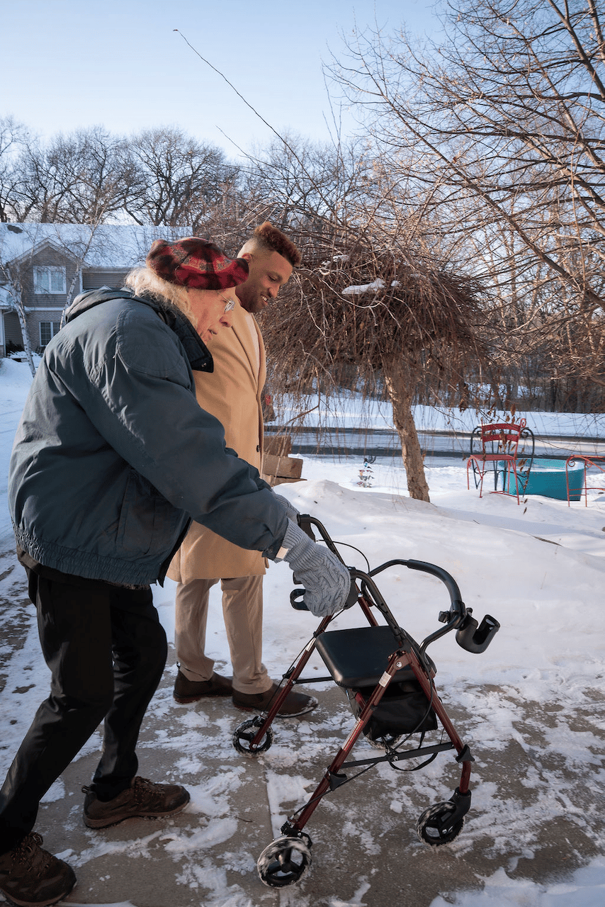 A caregiver assists a senior man using a walker on a snowy sidewalk during winter.