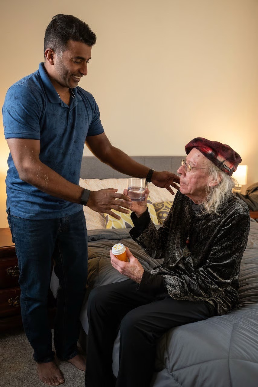 A smiling male caregiver hands a glass of water to an elderly man holding a bottle of medication