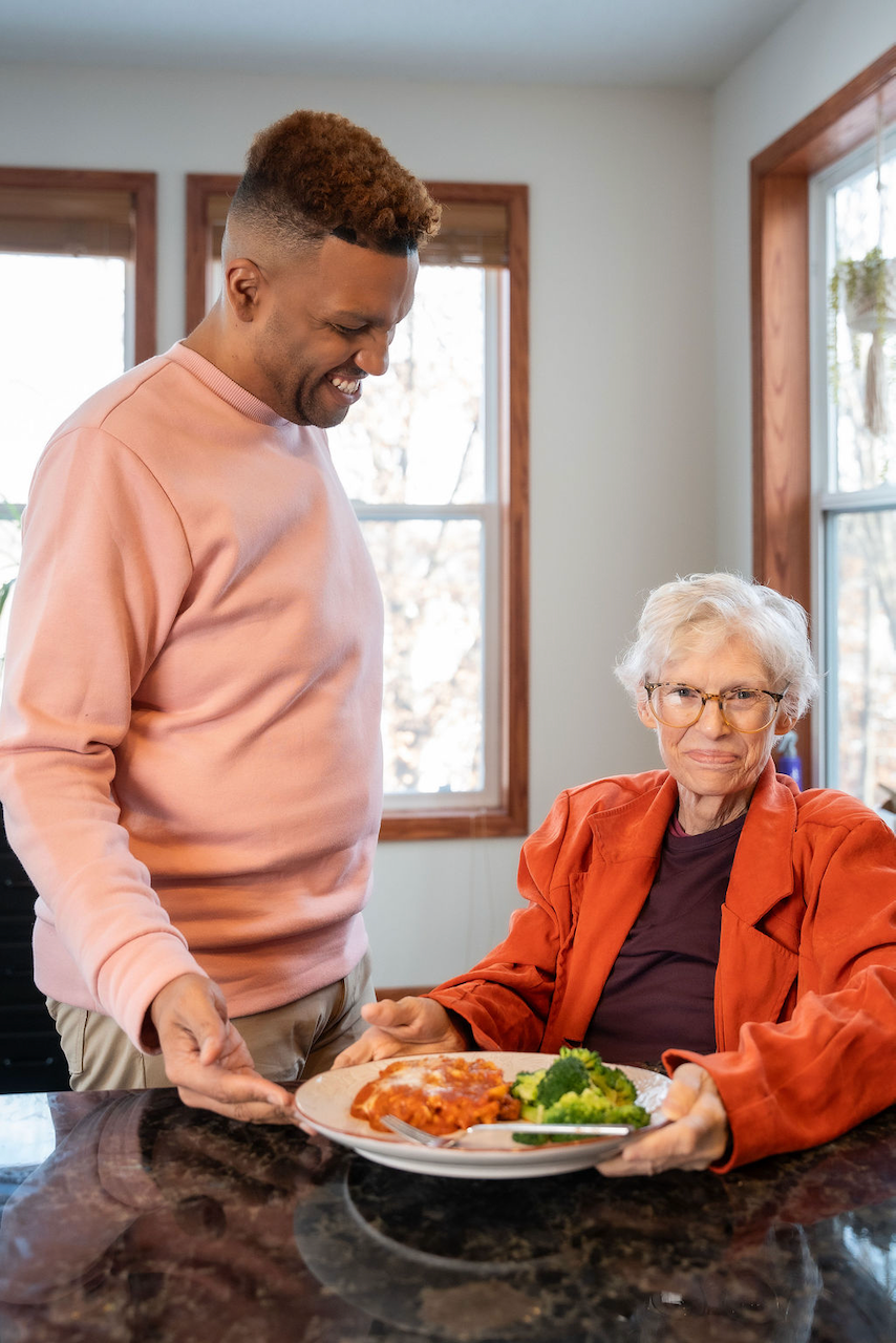 A smiling male caregiver serves a healthy meal of chicken and broccoli to a senior woman at home.