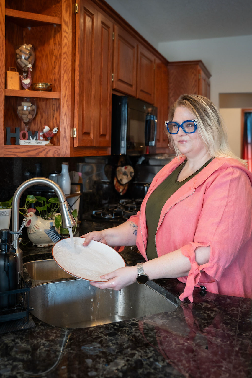 A blonde woman in a pink jacket washes a white plate in a stainless steel kitchen sink.