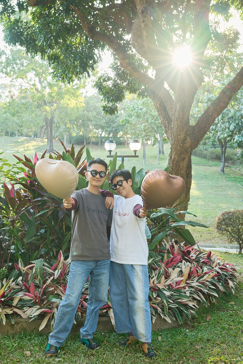 A happy couple wearing sunglasses holds heart-shaped balloons in a sunny park at sunset.