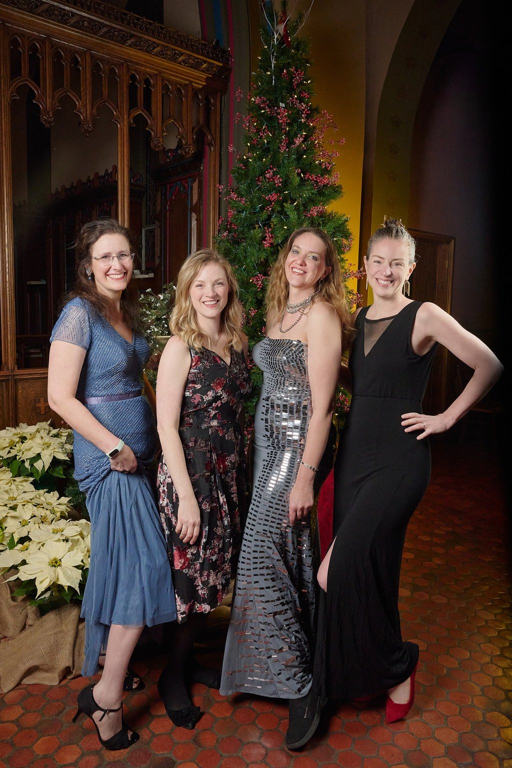 Four women in elegant evening gowns pose together in front of a decorated Christmas tree.