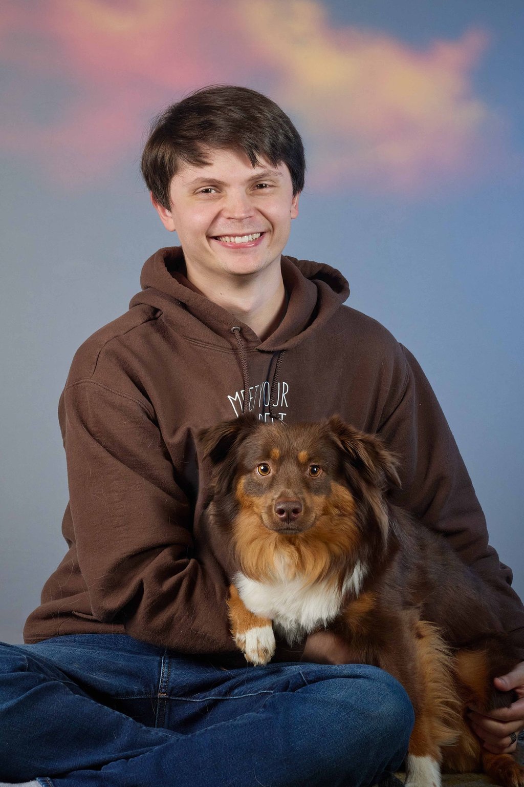 A smiling man in a brown hoodie sitting and holding his brown and white Australian Shepherd dog.