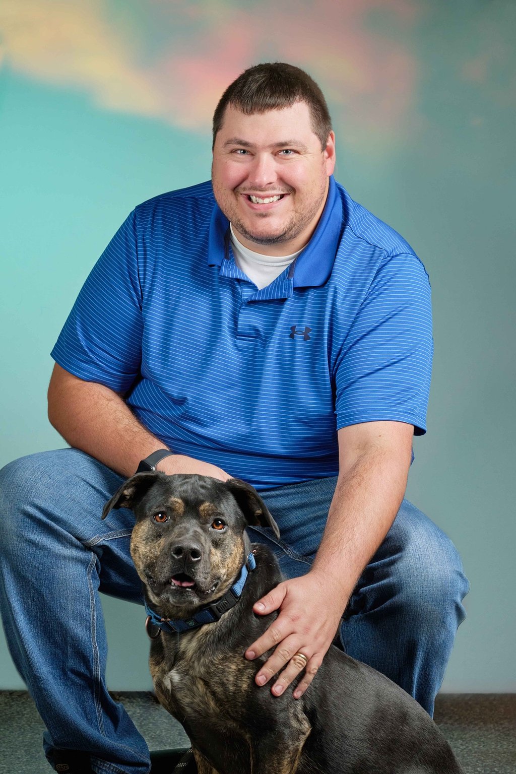 A smiling man in a blue polo shirt posing for a portrait with his brindle pit bull mix dog.