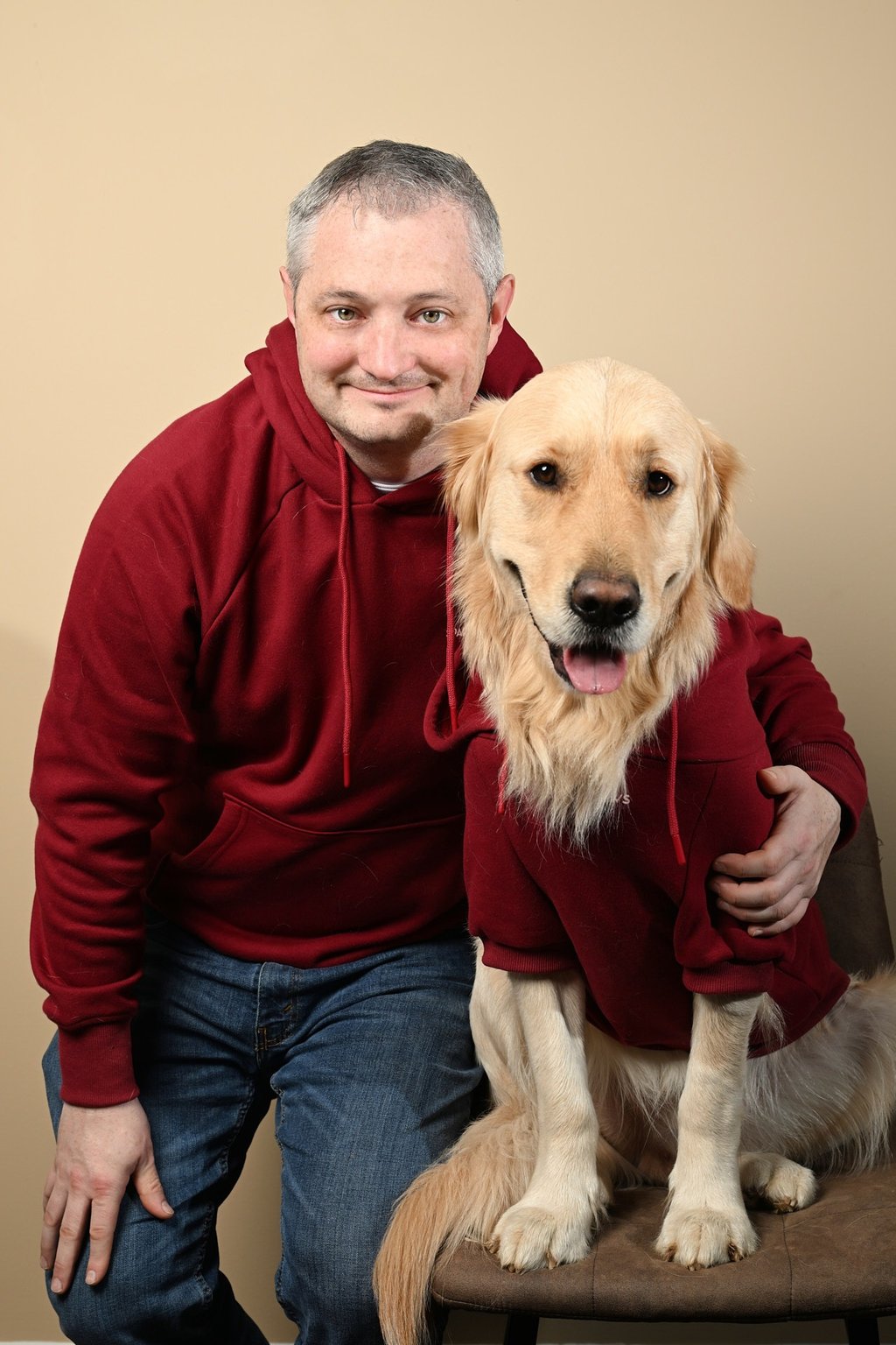 A smiling man and his Golden Retriever wearing matching maroon hoodies in a studio portrait.