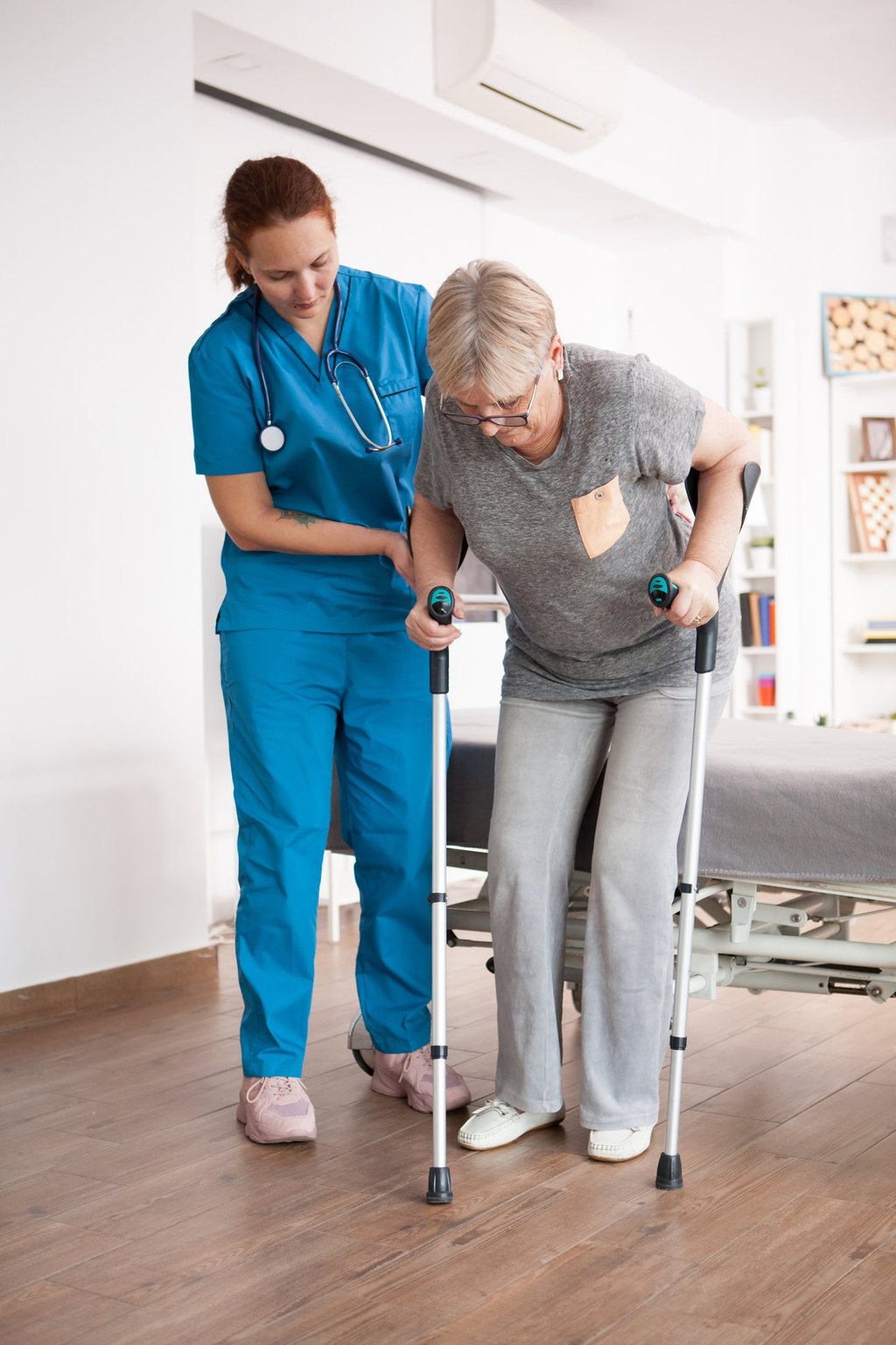 A female nurse in blue scrubs assisting an elderly woman using forearm crutches during physical therapy.