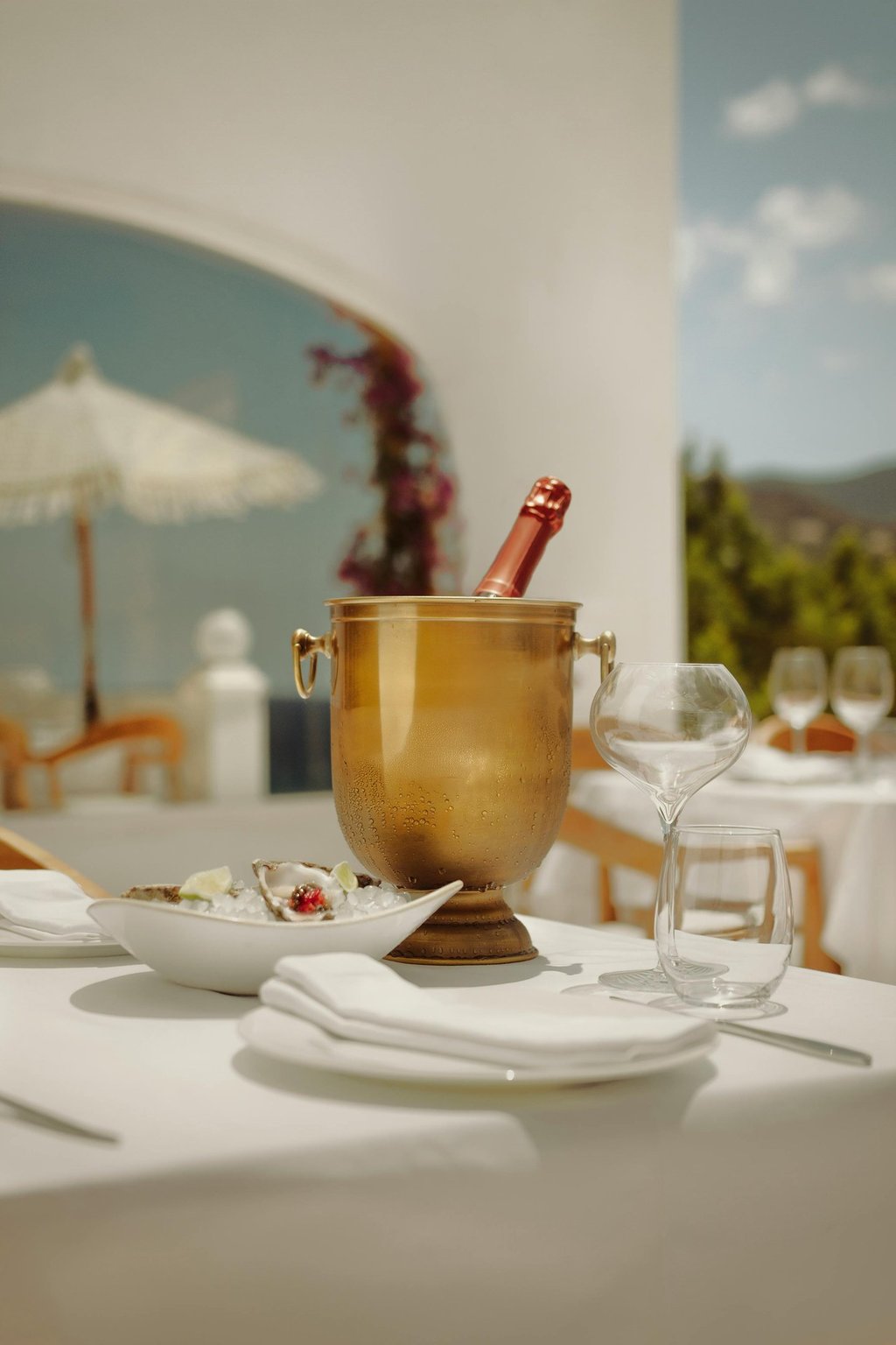 Outdoor dining table with champagne in an ice bucket, oysters on a plate, and empty glasses.
