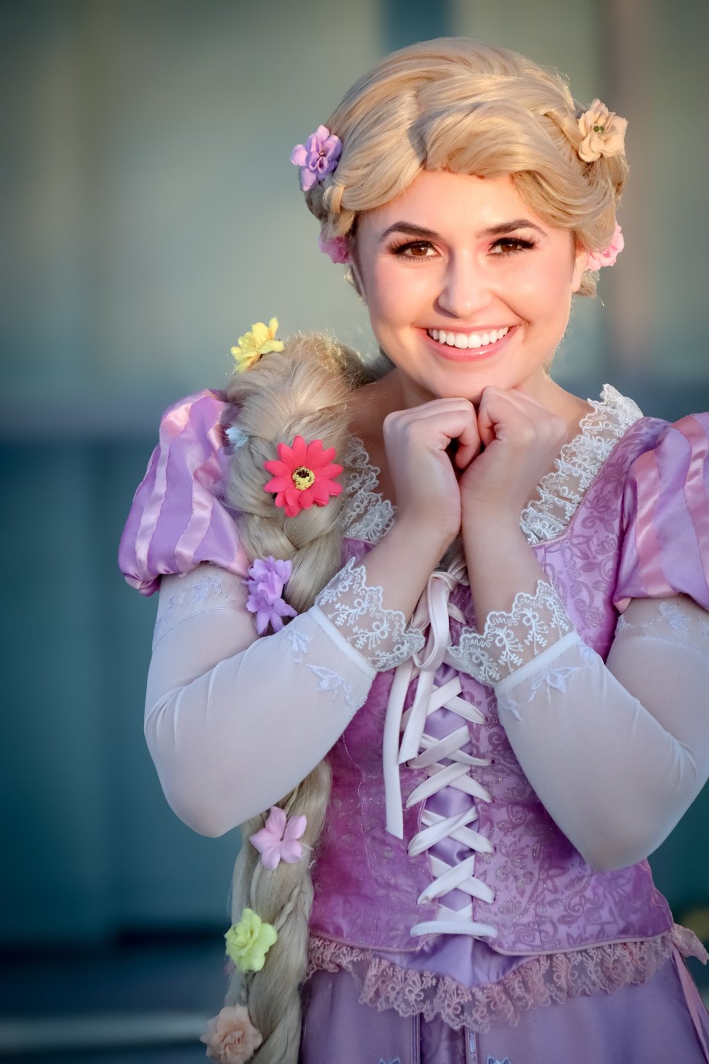 Smiling Rapunzel cosplayer wearing a purple floral dress with long braided blonde hair and flowers.