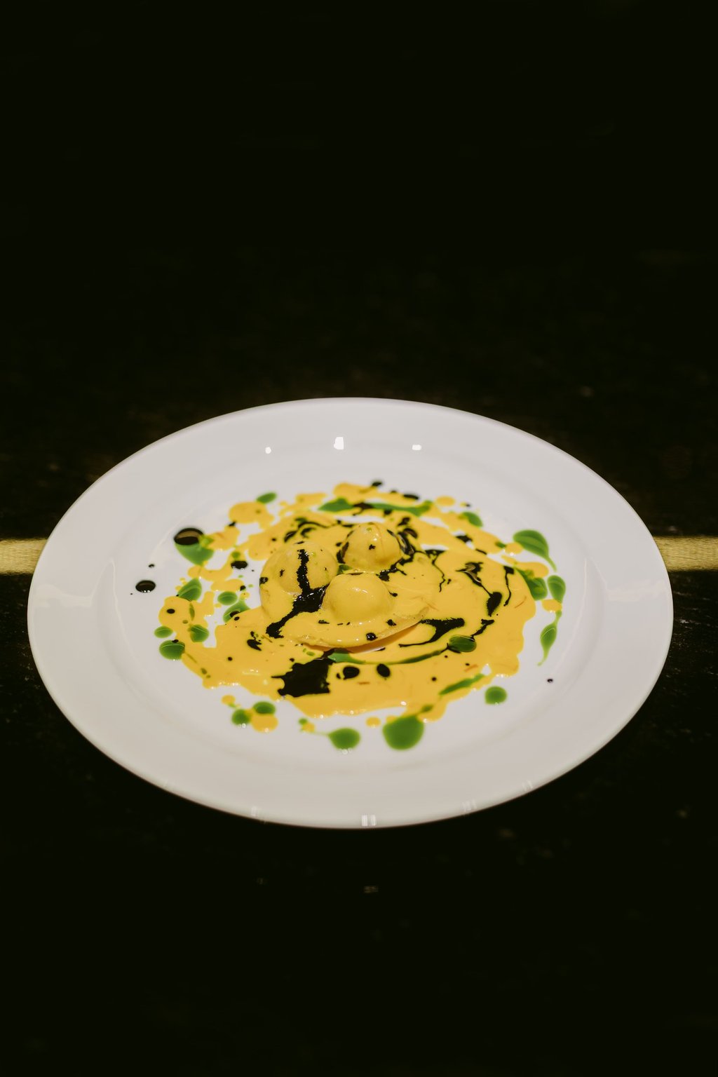 Raviolo signature dello chef Riccardo Quadri con salsa gialla e decorazioni verdi e nere.