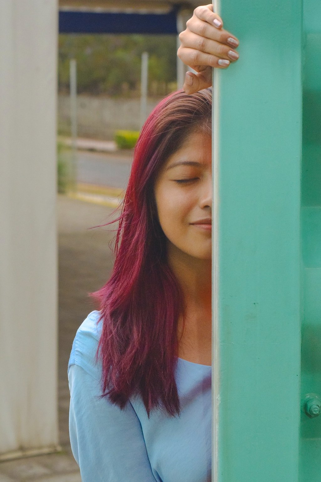Smiling woman with dyed magenta hair peeking from behind a teal pillar in an outdoor setting.