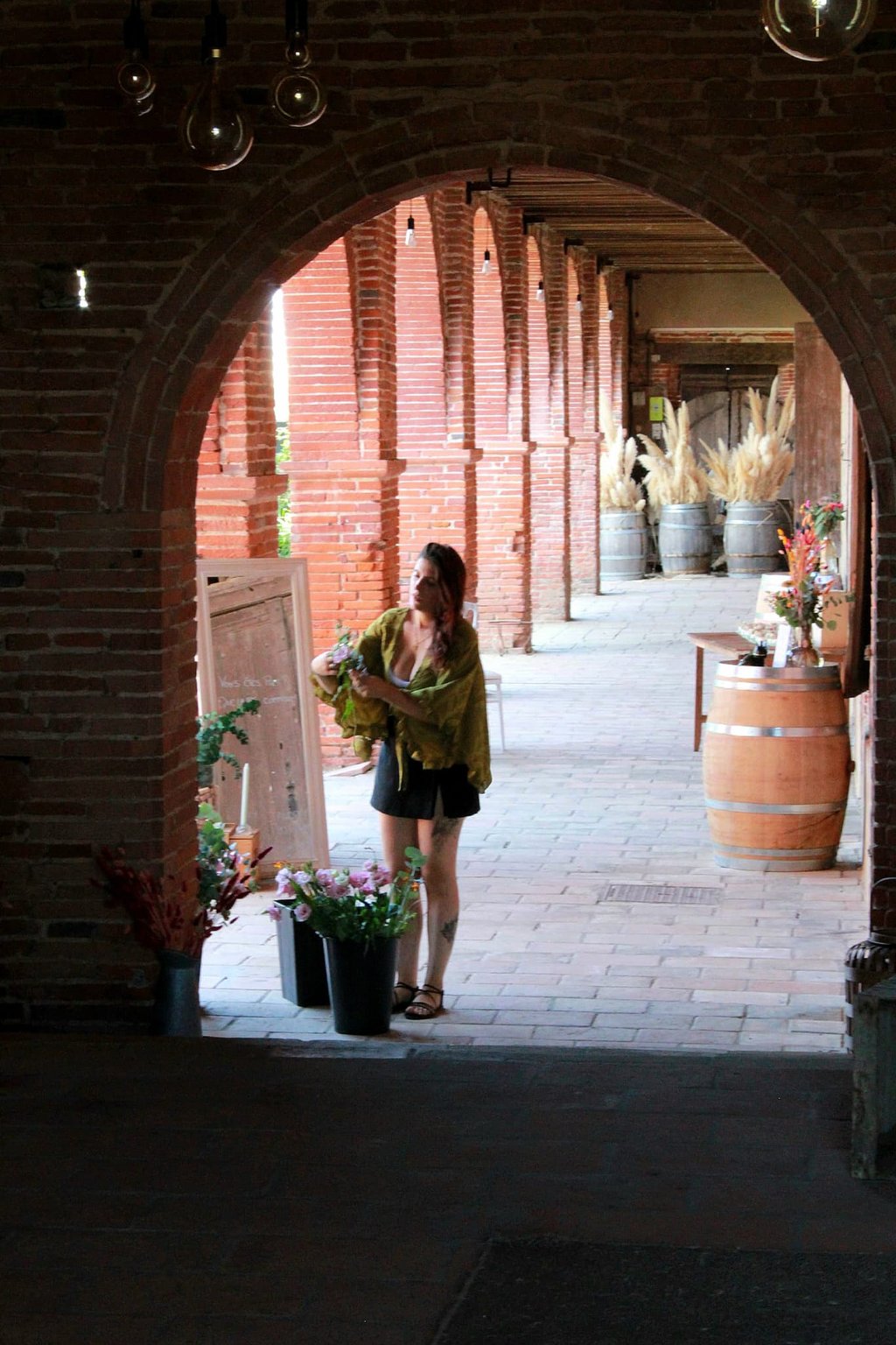 Estelle arrangeant des fleurs séchées dans des seaux sous une galerie en briques avec des arches
