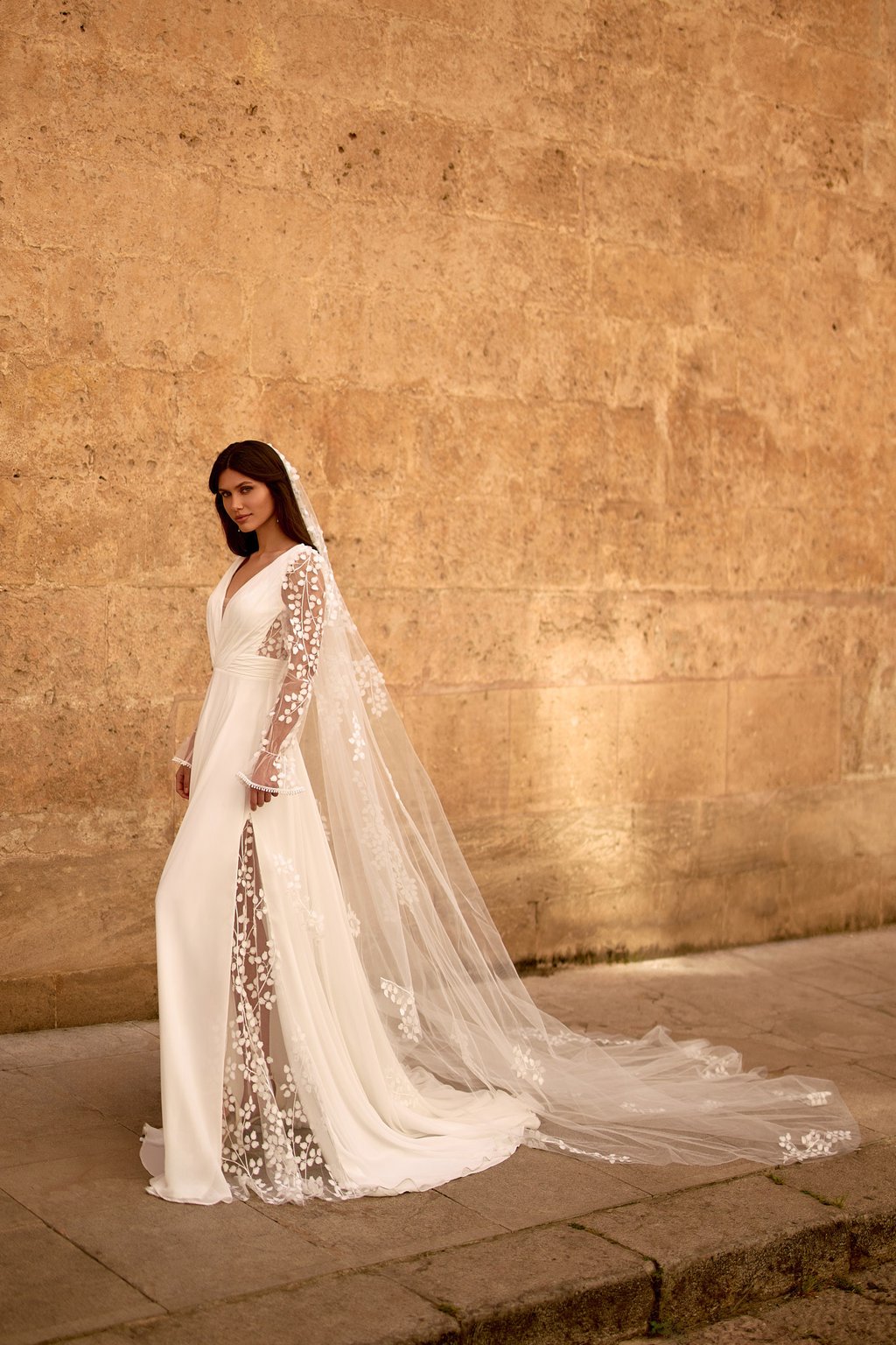 Elegant bride wearing a long sleeve lace wedding dress with a floor-length floral veil against a stone wall.