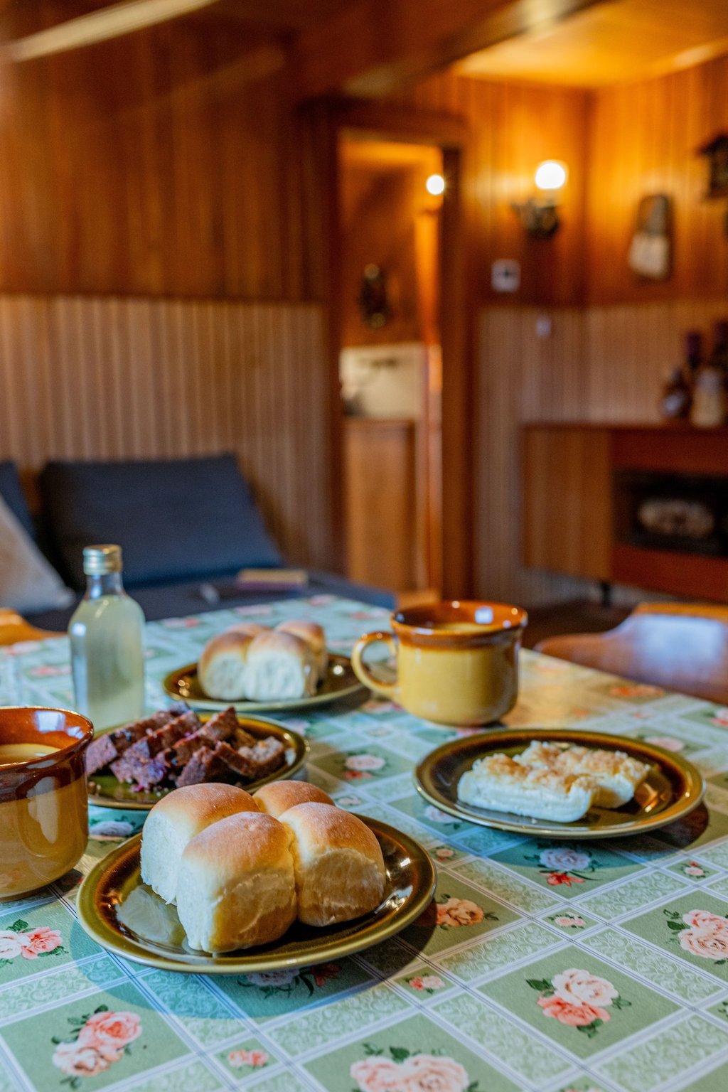 Homemade dinner rolls and pastries served on a floral tablecloth in a rustic wood-paneled dining room.