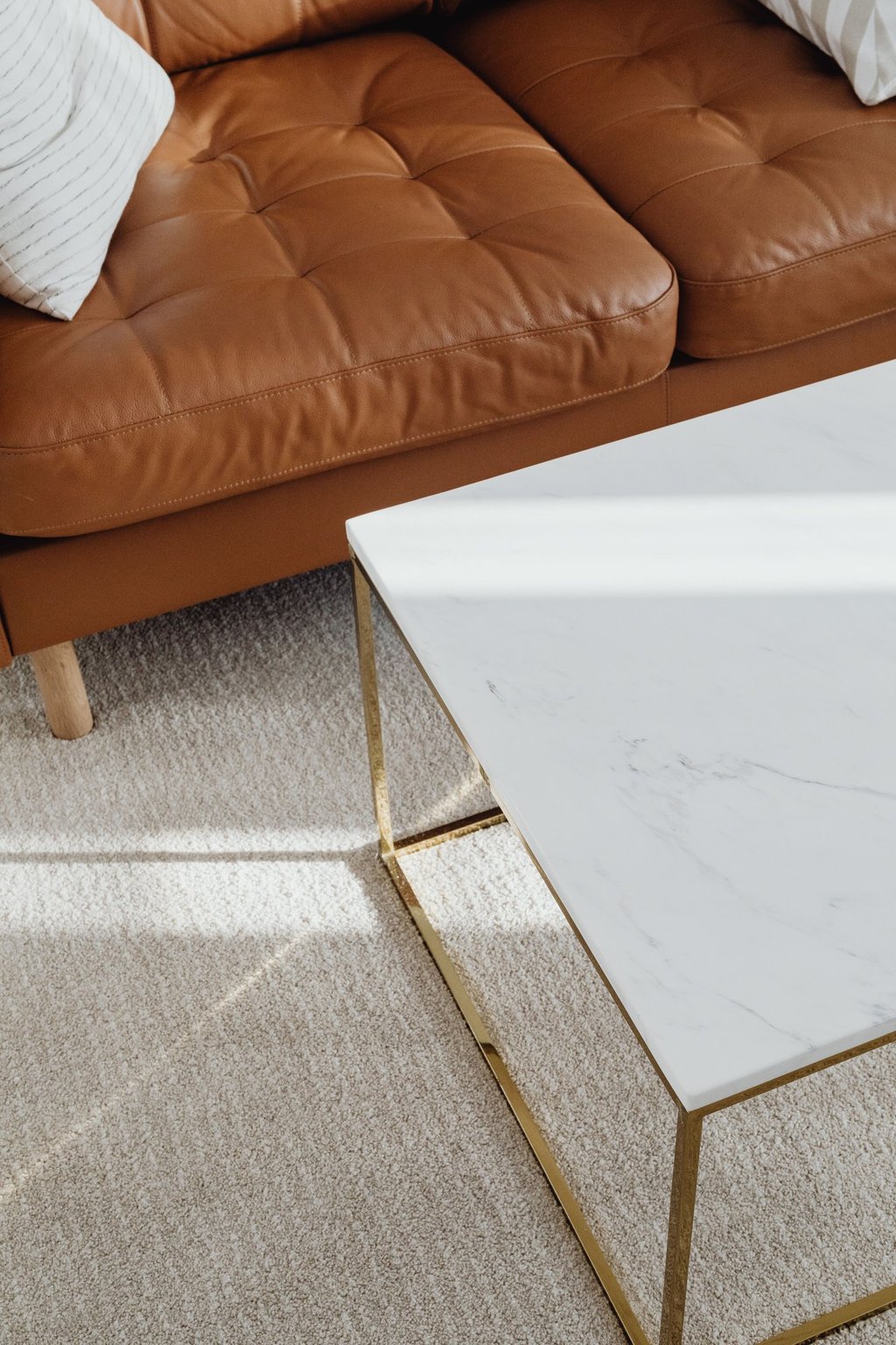 Modern white marble coffee table with gold legs sitting on a plush rug next to a tan leather sofa.