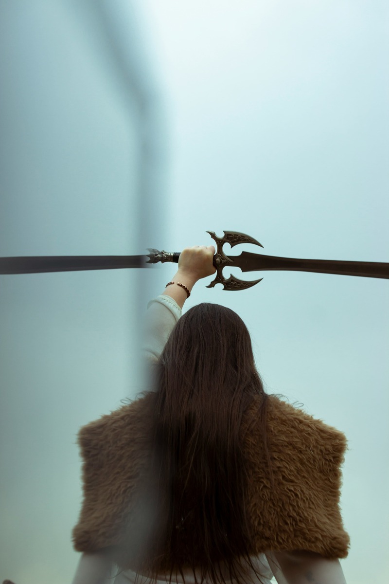 A woman in a fur vest holding a medieval fantasy sword against a foggy outdoor background.