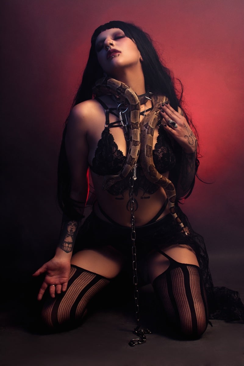 A gothic woman in black lace lingerie posing with a large boa constrictor snake in a red hazy studio.