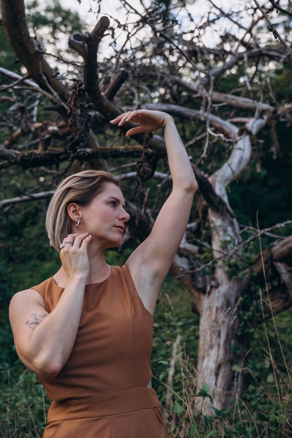 A blonde woman in a brown dress poses gracefully with raised arms in front of an old gnarled tree.