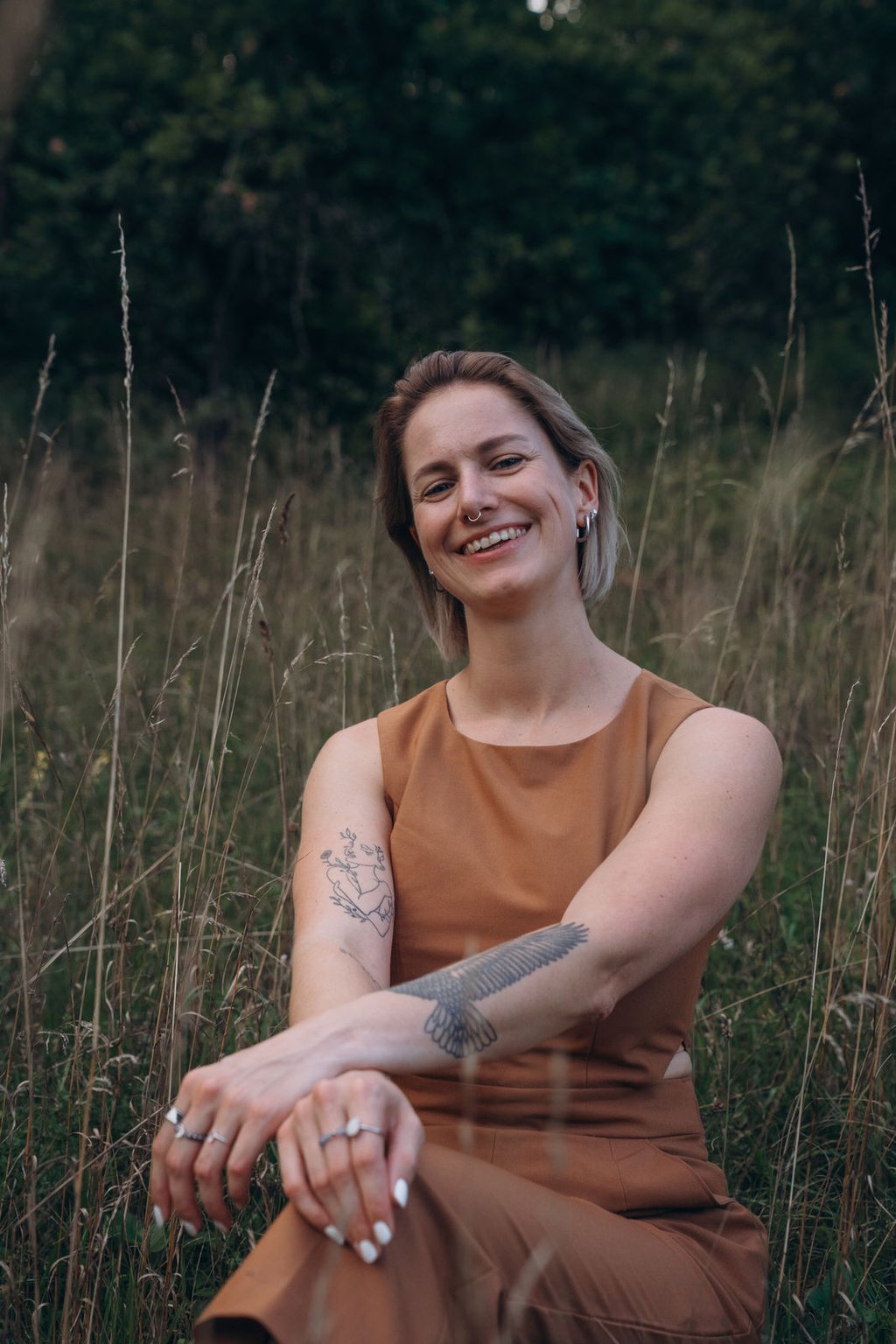 Smiling woman with tattoos wearing a brown dress sitting in a field of tall grass.