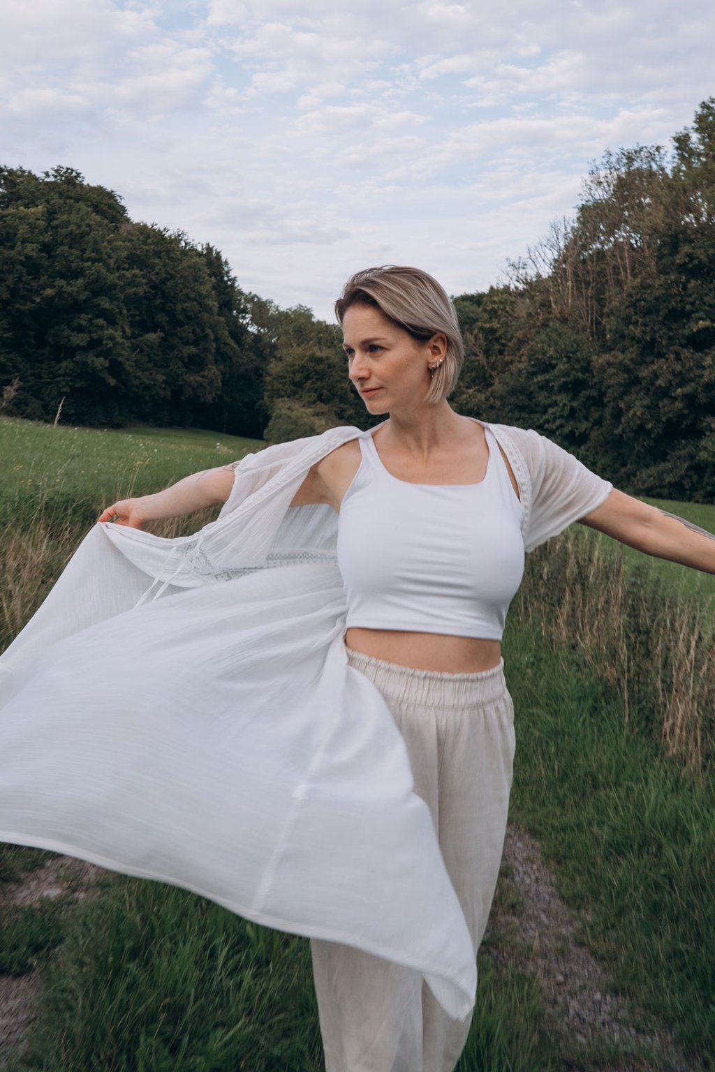A blonde woman in white boho clothing poses in a green meadow field with trees in the background.