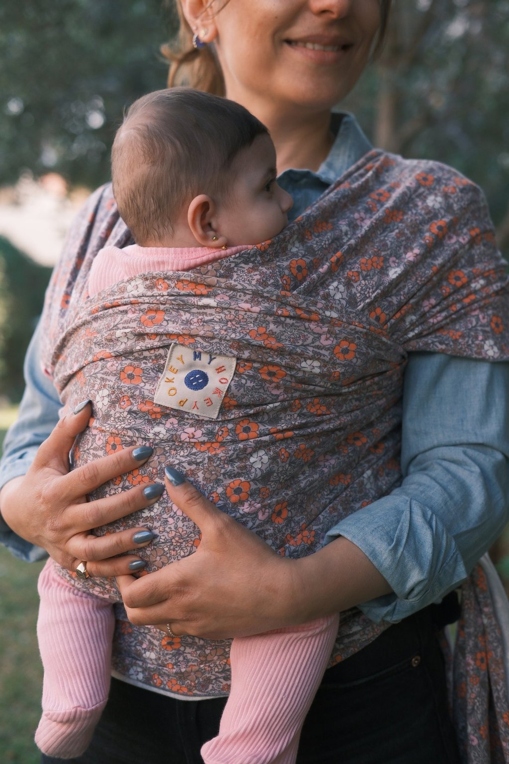 Mother holding her baby in a floral patterned ergonomic baby wrap carrier outdoors.