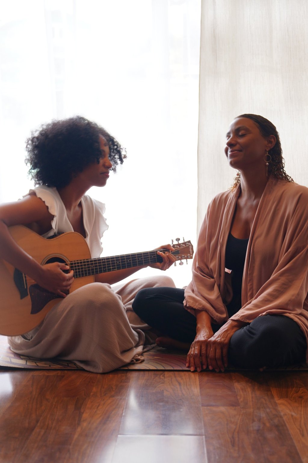 two women in front of a bright window sitting on a carpet playing a guitar.