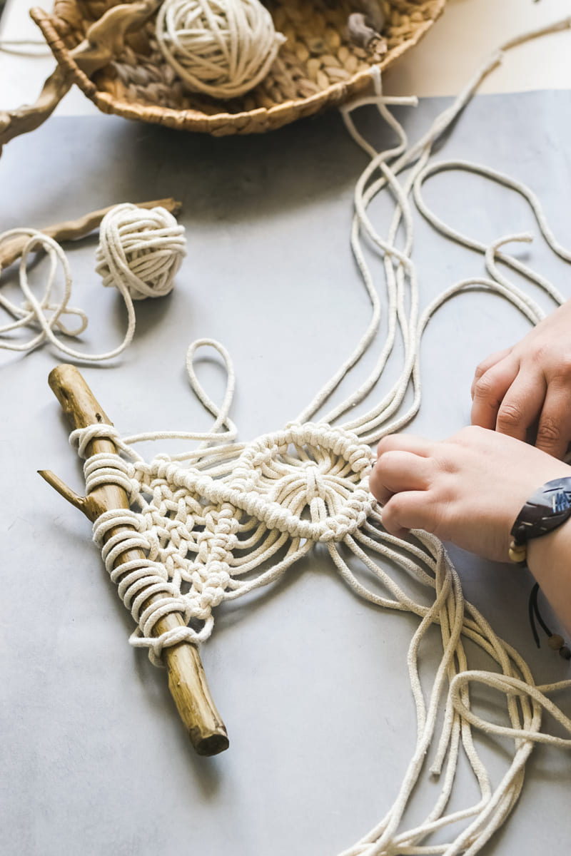 Manos elaborando un colgante de pared de macramé DIY usando cordón de algodón blanco con una rama de madera.