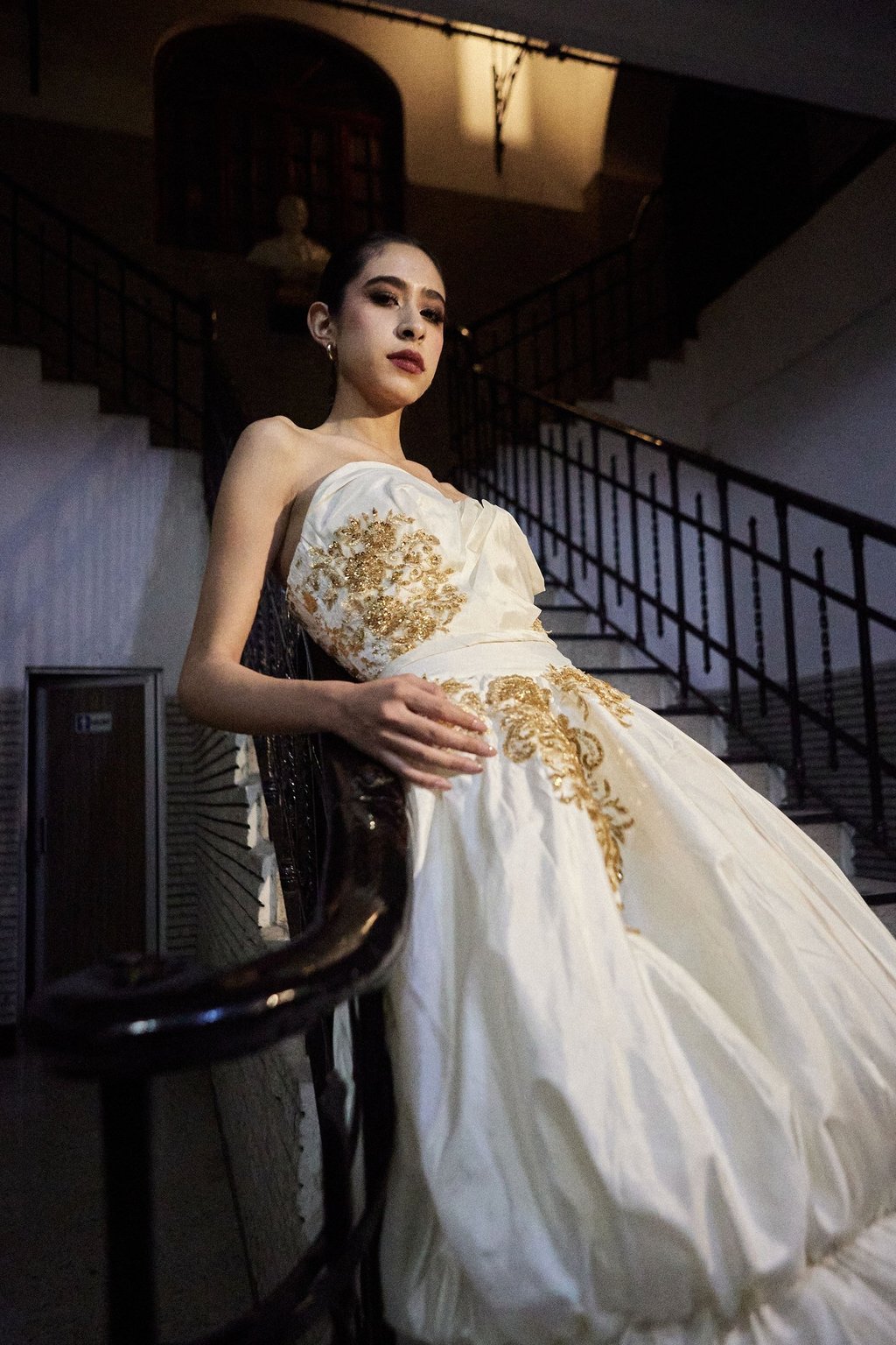 Elegant model in a white strapless gown with gold embroidery posing on a vintage staircase.