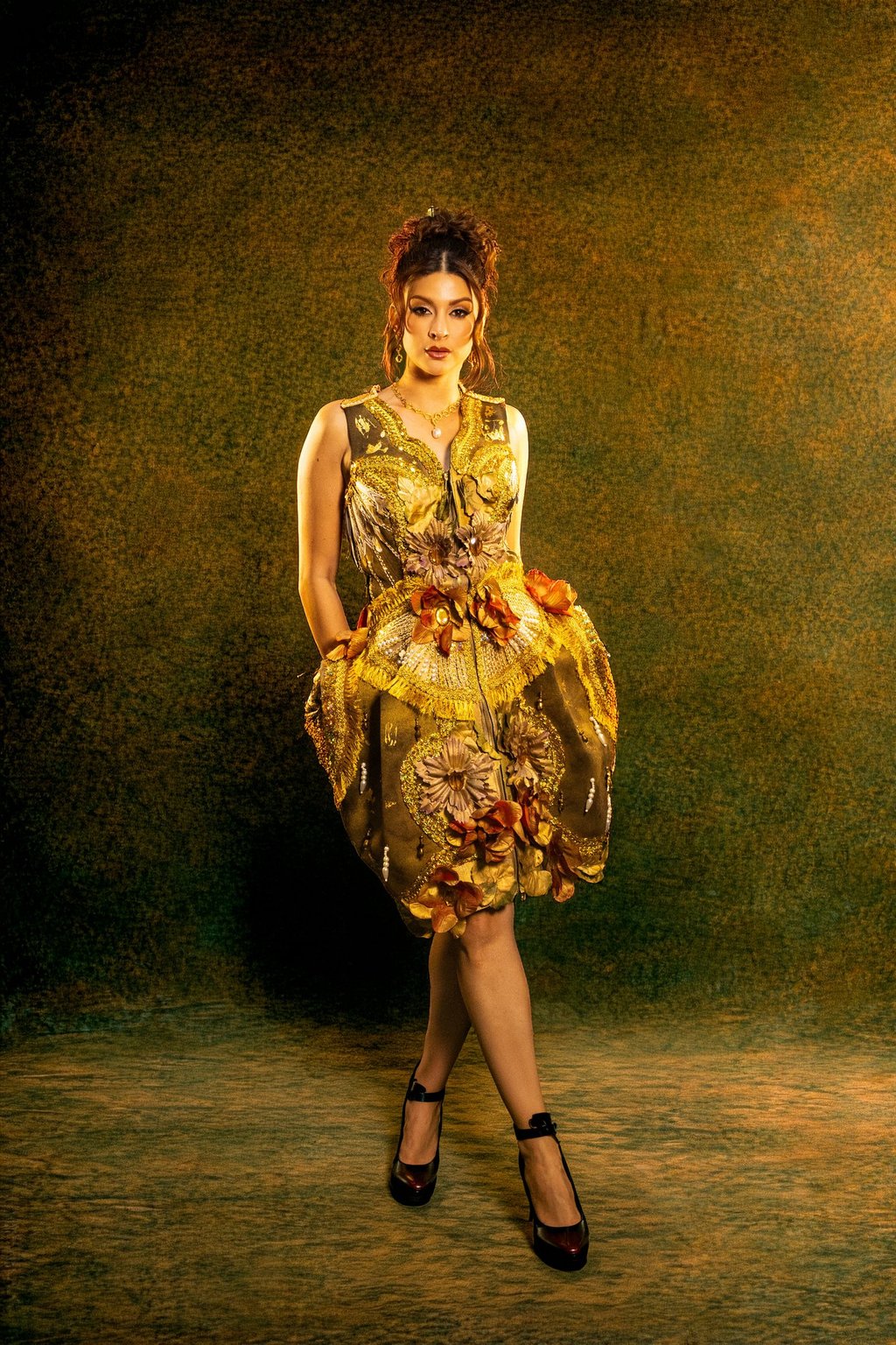 A woman poses in a structured gold couture dress with 3D floral accents and embroidery.