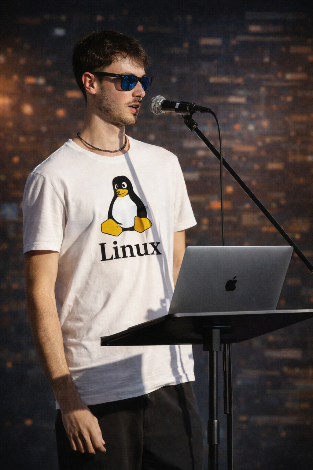 A man in a Linux penguin t-shirt giving a presentation with a MacBook and microphone.
