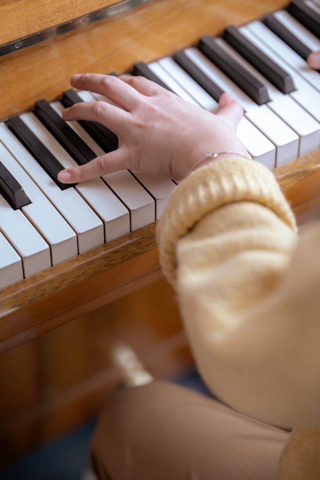 A person wearing a yellow sweater plays a wooden upright piano during a music lesson.