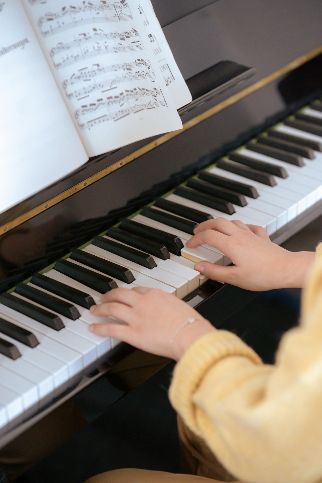 A student's hands playing classical music on a black upright piano with sheet music displayed.