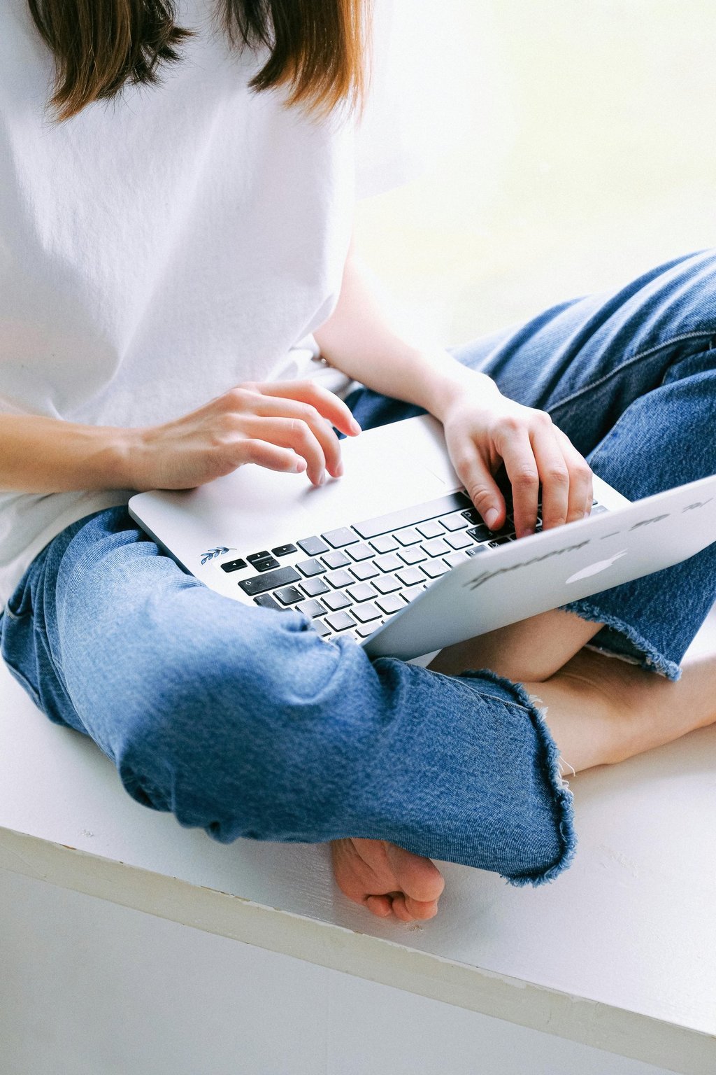 A person in blue jeans sitting cross-legged while typing on a laptop computer.