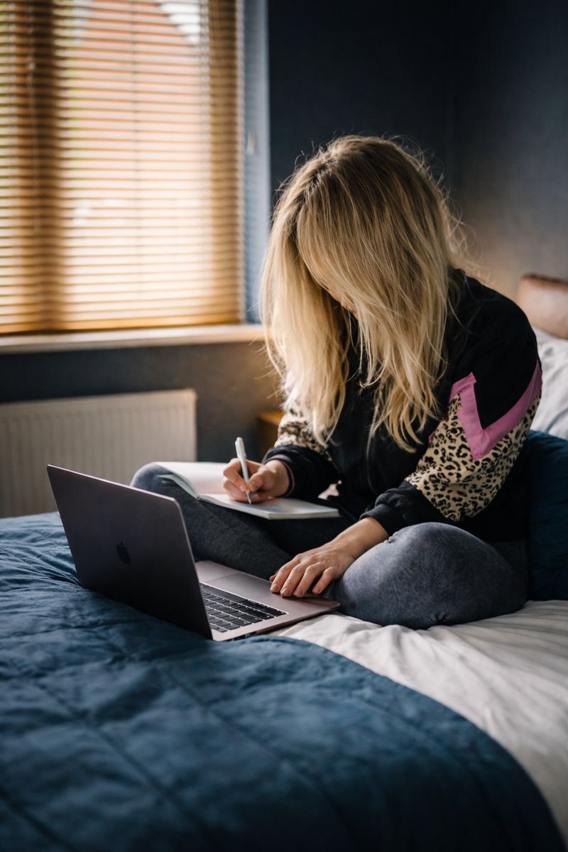 A young woman sits on her bed, using a laptop and notepad
