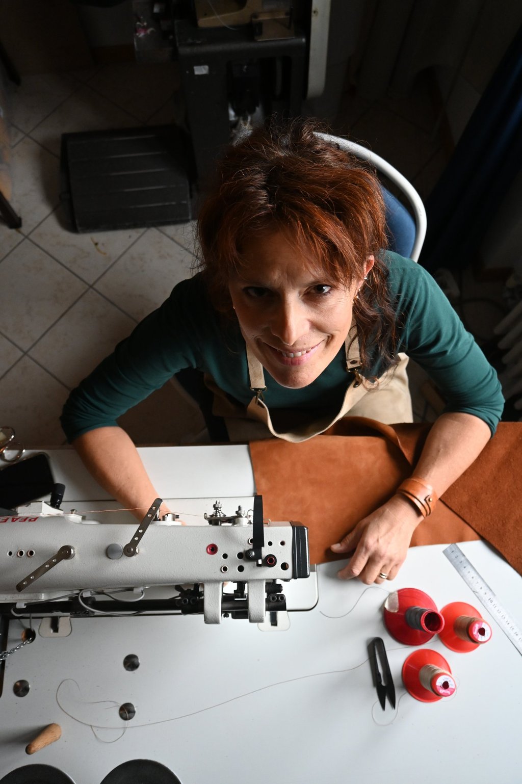 Female leather artisan smiling at her industrial sewing machine while crafting a handmade leather bag.