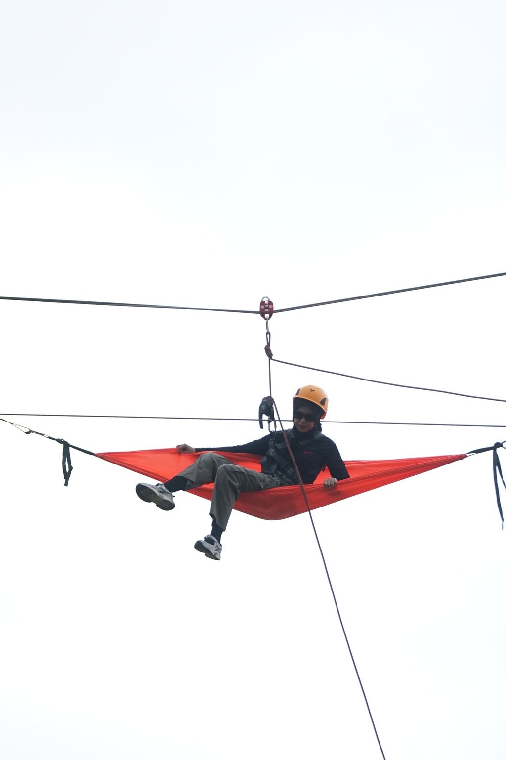 A person wearing a safety helmet relaxes in an orange hammock suspended high on a zip line cable.