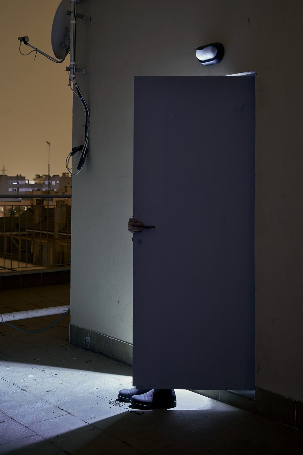 A person opening a door on a dark rooftop balcony with an outdoor light and city skyline.