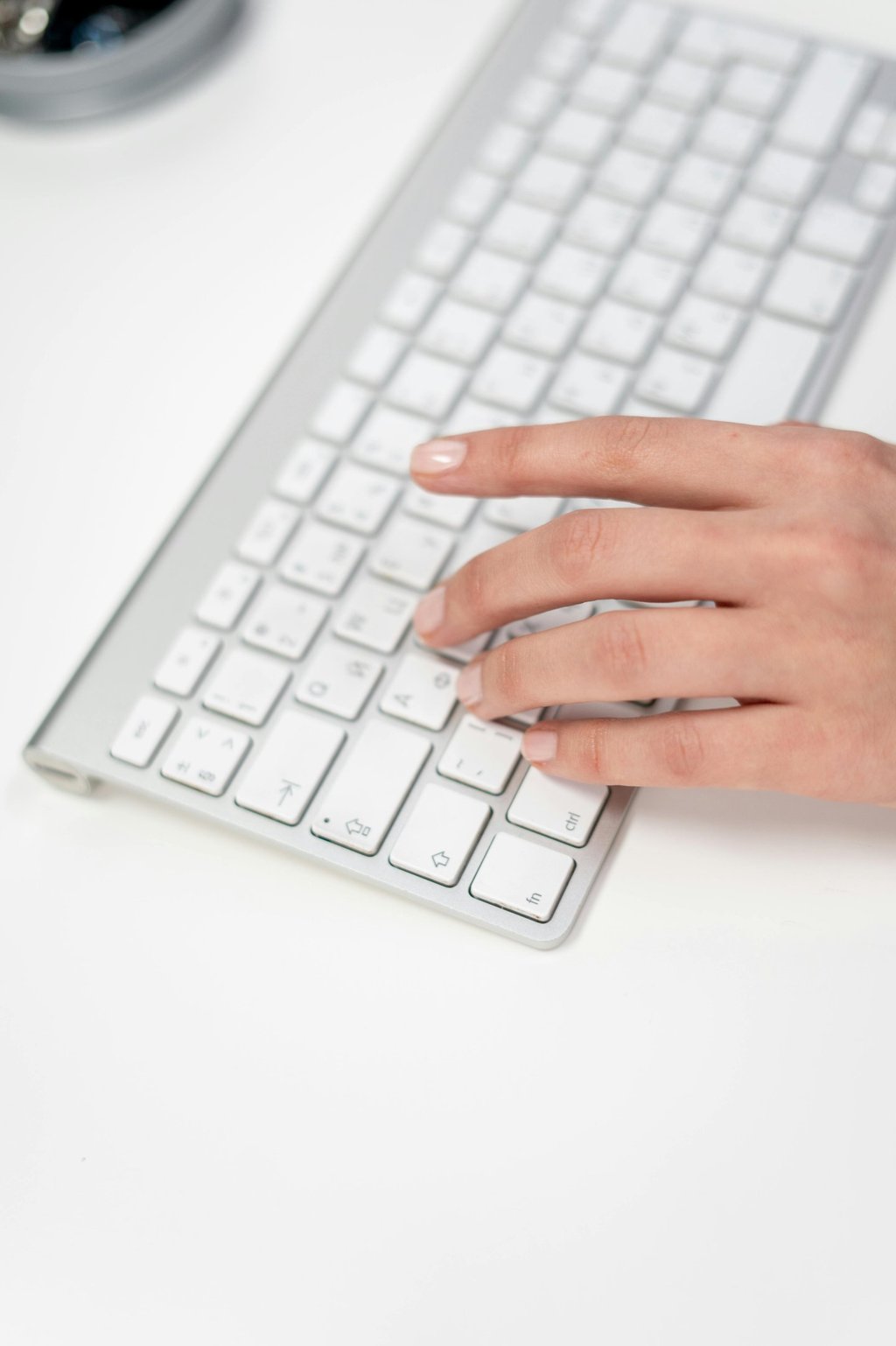 A person's hand typing on a slim silver and white wireless computer keyboard on a white desk.