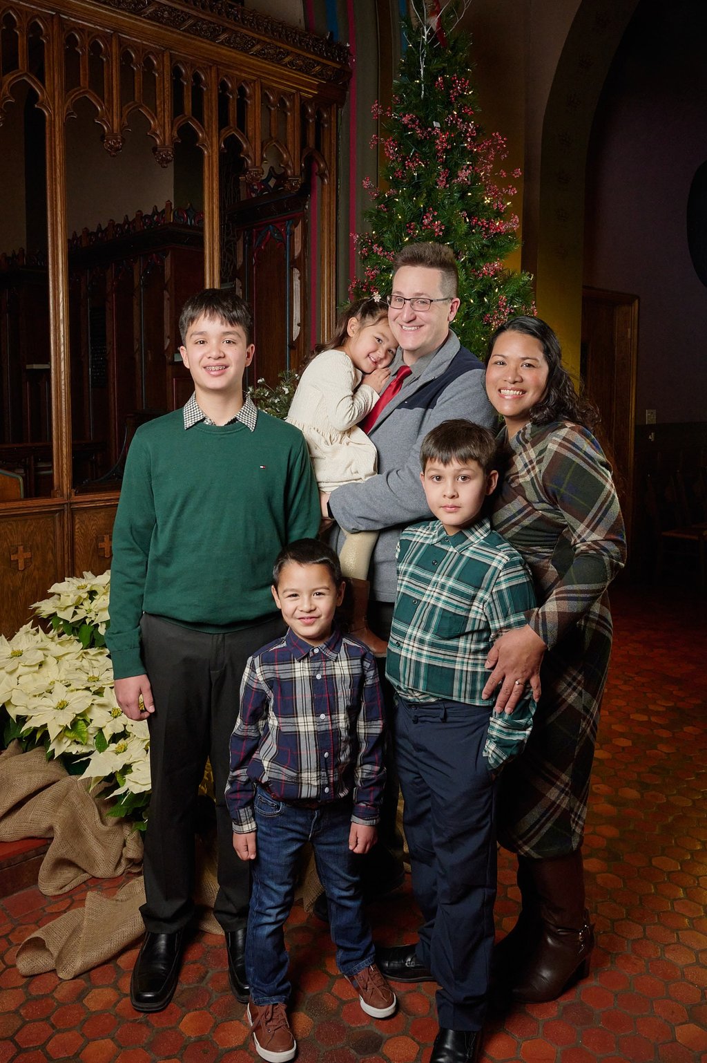 A happy family posing for a Christmas portrait in a church with a decorated tree and poinsettias.