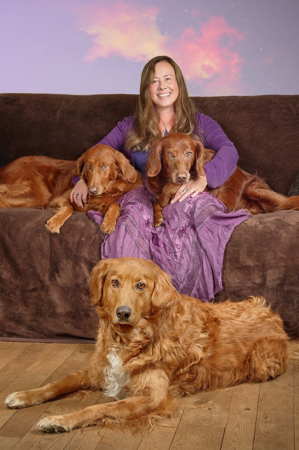 Smiling woman in purple sitting on a brown sofa with three golden Irish Setter dogs.