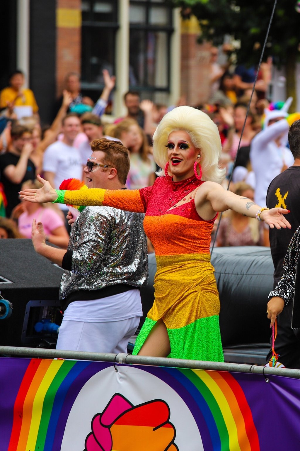 A drag queen in a rainbow dress with large blonde wig holds her arms out in greeting