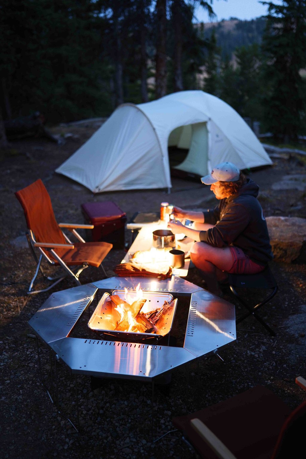 a man sitting at a table with a plate of food next to a campfire