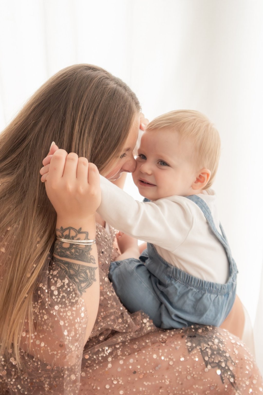 A smiling mother with a forearm tattoo holding her blonde toddler boy in blue overalls.