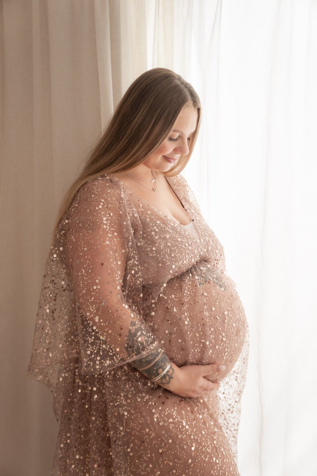 A smiling pregnant woman with tattoos wears a rose gold sequin maternity dress for a studio photo shoot.