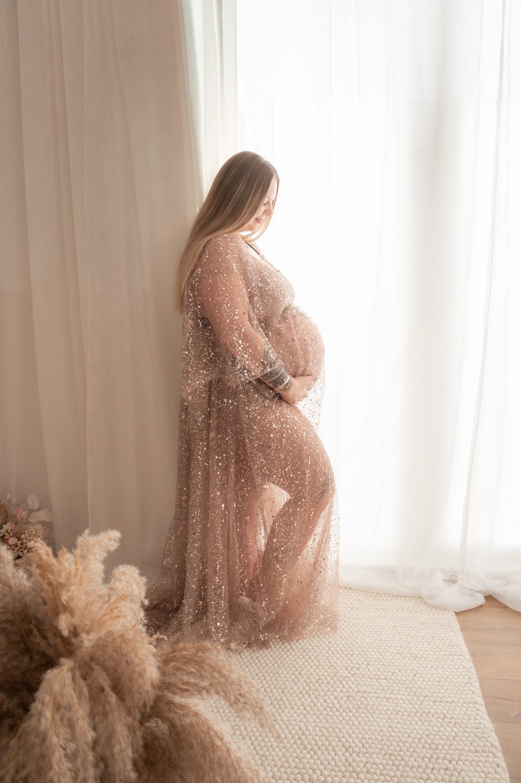 Pregnant woman in a sparkly maternity gown posing for a photoshoot by a window.